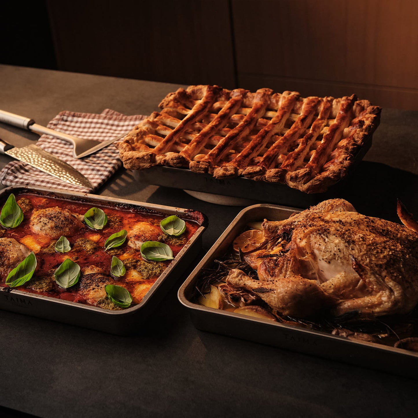 Three roasted dishes on a dark surface with a checkered cloth and utensils.
